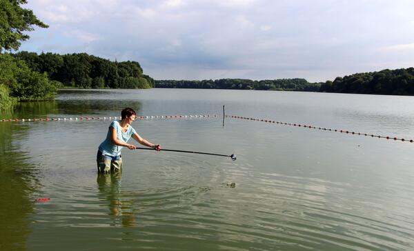 Eine Frau steht in einem See und entnimmt eine Wasserprobe. Dafür hält sie einen langen Stab, an dessen Ende das Probengefäß angebracht ist.