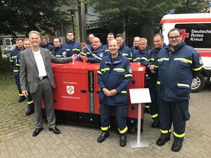 Viele Einsatzkräfte des Technischen Hilfswerks mit blauen Uniformen stehen mit Landrat Jan Peter Schröder neben einer Art roten Kiste.