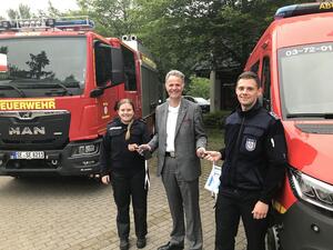 Eine Frau und ein Mann in dunkelblauer Einsatzkleidung stehen gemeinsam mit Landrat Jan Peter Schröder vor zwei großen Einsatzwagen der Feuerwehr.