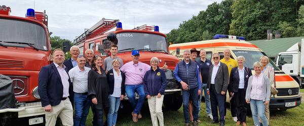 Vor zwei alten Feuerwehrlöschfahrzeugen und einem Krankenwagen stehen Frauen und Männer und lachen in die Kamera.