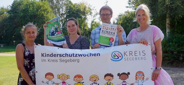 Drei Frauen und ein Mann halten ein Plakat zum Thema Kinderschutzwochen im Kreis Segeberg in den Händen. Sie stehen im Freien auf einer Wiese.